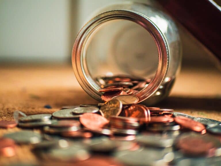 Des pièces de monnaie s'échappant d'un bocal en verre renversé sur une surface, avec un mélange de pièces de monnaie et d'autres pièces de monnaie de couleurs cuivre et argent.